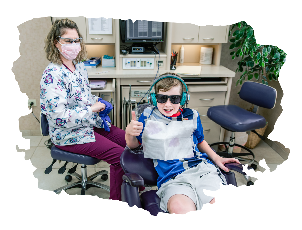 A young patient wearing sunlasses and headphone gives a thumbs up wile sitting in the dental chair.